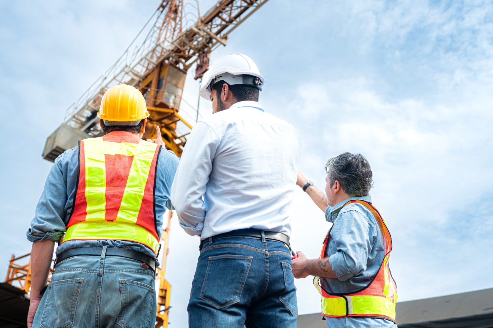 Drei Personen stehen vor einem Kran auf einer Baustelle und bekommen eine Unterweisung. 